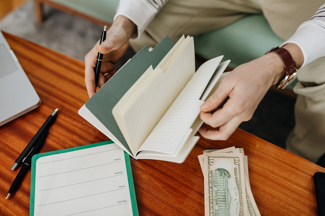About A person sits at a wooden table organizing notes in a notebook and handling cash.